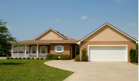 A single-story house with a two-car garage and a welcoming porch, set in a beautiful landscape with a well-manicured lawn, neat shrubs, and a clear blue sky.