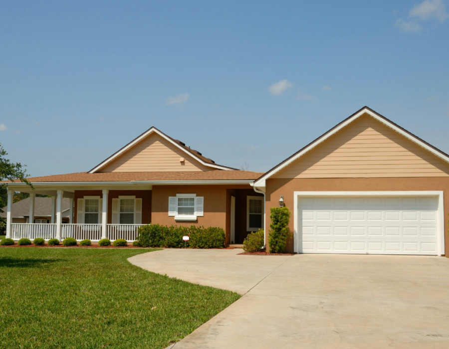 A single-story house with a two-car garage and a welcoming porch, set in a beautiful landscape with a well-manicured lawn, neat shrubs, and a clear blue sky.